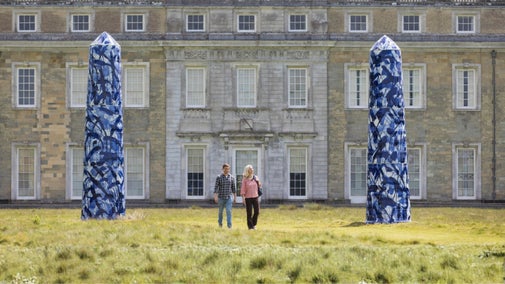 Two visitors walking past giant blue and white ceramic monoliths from the Expressions in Blue exhibition, in Petworth House's parkland with house in background.